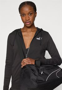 Μαύρο φούτερ με φερμουάρ και λογότυπο στο στήθος, συνδυασμένο με μαύρη τσάντα που έχει μια λευκή διαγώνια διακοσμητική λεπτομέρεια και φερμουάρ.