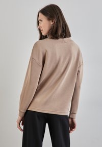 Felpa marrone chiaro con vestibilità rilassata, scollatura rotonda e maniche lunghe. Il tessuto appare morbido e liscio con dettagli di cucitura minimi.