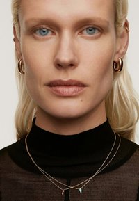 Pendientes de aro en oro rosa y collares de oro en capas con piedras de acento coloridas, sobre un top negro de cuello alto. Piel suave y cabello rubio lacio.