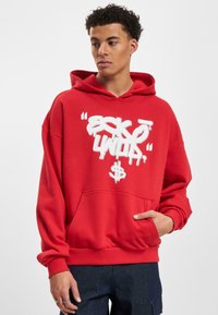 Felpa con cappuccio rossa realizzata in tessuto morbido, caratterizzata da una grande grafica bianca in stile graffiti e da una tasca a marsupio frontale.