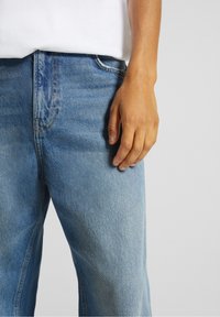 Jeans de mezclilla azul claro con corte recto, presentan costuras visibles y un diseño clásico de cinco bolsillos, combinados con una camisa blanca.