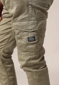 Khaki cargobroek met een textuurstof, meerdere zakken en een zwart label aan de zijkant, wat een functioneel en duurzaam ontwerp benadrukt.