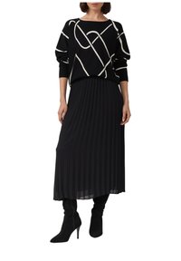 Czarny sweter z kremowymi, ukośnymi liniami, nałożony na plisowaną czarną spódnicę. Modelka ma na sobie czarne botki na wysokim obcasie sięgające do kolan.