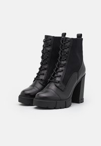 Bottines noires à talons hauts en cuir et matériau tricoté, dotées d'un laçage sur le devant, d'une semelle épaisse et d'un talon bloc haut distinct.