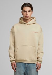 Beige hættetrøje med lomme foran og ribbede manchetter. Har "Slow Sunday Club" broderet i orange på brystet.