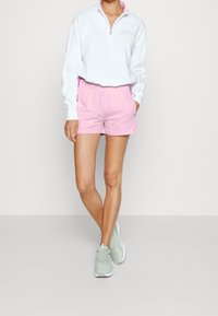 Persona vestida con un sudadera blanca de cremallera de cuarto, pantalones cortos rosas y zapatillas verde claro, de pie con una pierna cruzada sobre la otra.