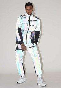 Giacca iridescente con dettagli neri, dotata di diverse tasche frontali, abbinata a pantaloni iridescenti coordinati. Entrambi presentano un design slim fit.