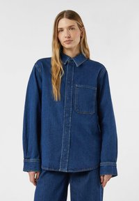 Koszula dżinsowa w ciemnoniebieskim kolorze z długimi rękawami, zapięciem na guziki z przodu i kieszenią na piersi. Posiada ostro zakończony kołnierz oraz luźny krój. Gładka faktura.