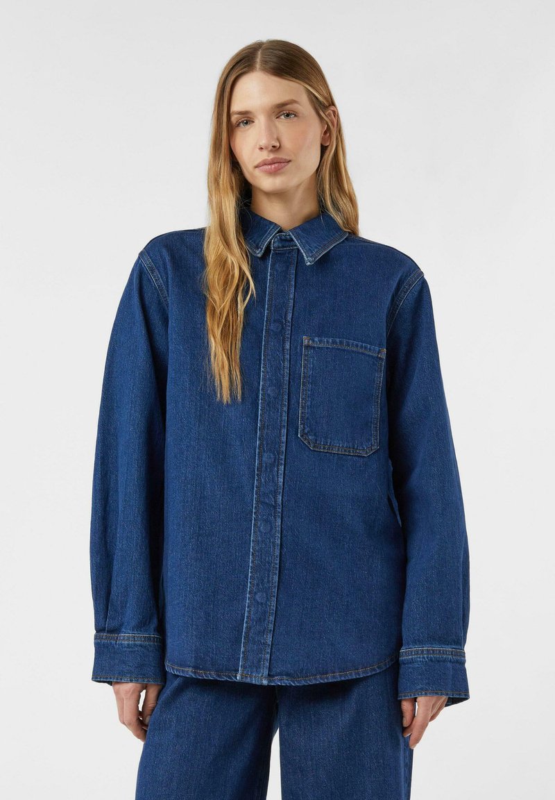 Koszula dżinsowa w ciemnoniebieskim kolorze z długimi rękawami, zapięciem na guziki z przodu i kieszenią na piersi. Posiada ostro zakończony kołnierz oraz luźny krój. Gładka faktura.