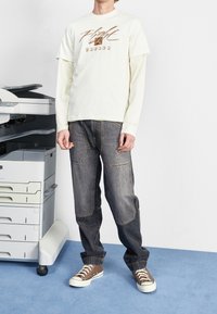 Valkoinen lyhythihainen t-paita, jossa on "Flight"-logo, kerrostettuna pitkähihaisen paidan päälle, yhdistettynä tummansinisiiin cargo-farkkuihin ja ruskeisiin lenkkareihin.