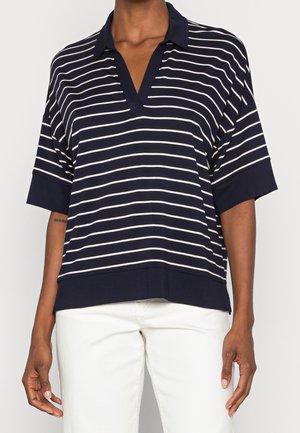 Mujer con una camiseta de manga corta de rayas horizontales en azul marino y blanco, con pantalones blancos, de pie frente a un fondo liso.