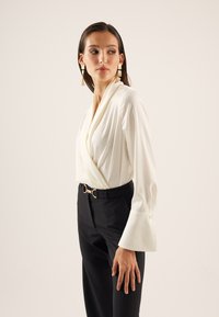 Blusa bianca avvolta con maniche lunghe e polsini abbottonati, abbinata a pantaloni neri sartoriali con una fibbia rotonda in tonalità oro.