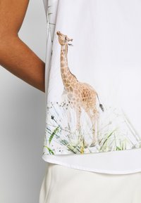 Personne portant une chemise blanche avec un imprimé girafe et herbe sur le côté, associée à un pantalon de couleur claire.