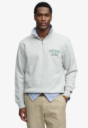 Homme portant un sweat-shirt gris clair à demi-fermeture éclair avec le texte "SUPERDRY JAPAN", une chemise bleue à col en dessous, un pantalon beige et une montre-bracelet.