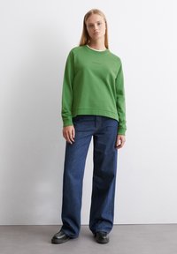 Grön sweatshirt med ribbade ärmar och fåll, i kombination med mörkblå vida jeans och svarta skor, mot en neutral bakgrund.