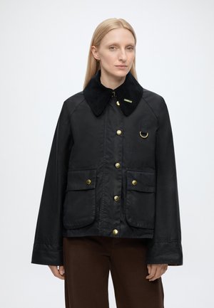 Frau mit glattem blondem Haar, die eine schwarze Jacke mit weitem Kragen, goldenen Knöpfen, zwei vorderen Taschen und braune Hosen vor einem schlichten Hintergrund trägt.