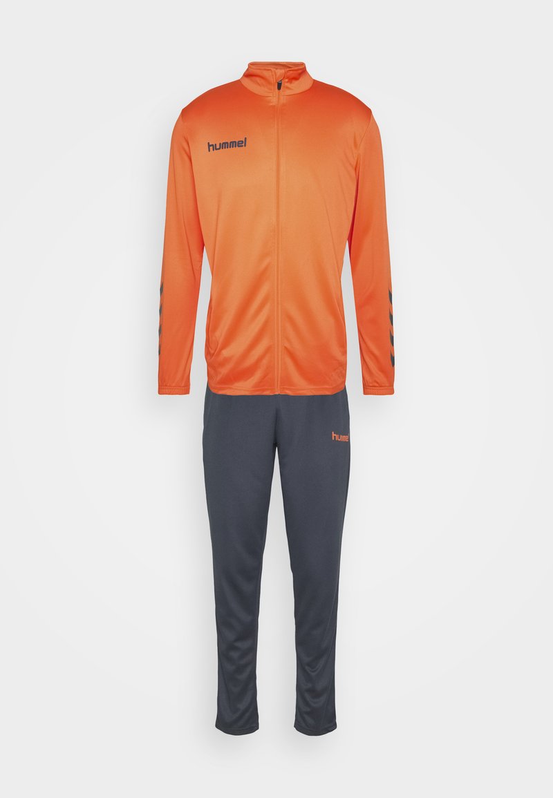 Pomarańczowa poliesterowa kurtka dresowa z wysokim kołnierzem, zamkiem błyskawicznym z przodu i czarnymi detalami. Połączona z szarymi spodniami z poliestru z kieszeniami po bokach.
