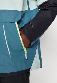 Casaco em bloco de cores com secções em azul claro, teal e azul marinho. Possui um fecho amarelo e detalhe do logótipo na manga. Textura suave.