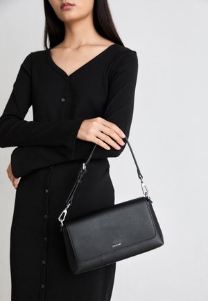 Černá kožená kabelka s texturovaným povrchem, odnímatelným popruhem a stříbrným kováním. Nese ji osoba v černých šatech s dlouhým rukávem.