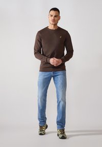 Brown long-sleeve sweatshirt with a small yellow logo, paired with light blue jeans and camouflage-patterned shoes. Casual fit and texture evident.