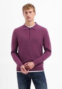 Paarse langwerpige polo met een ritssluiting bij de kraag, gemaakt van zachte stof. Draagt bij blauwe spijkerbroeken, met een effen ontwerp en aansluitende manchetten.