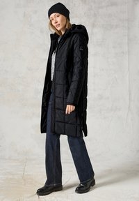 Svart vadderad lång kappa med huva, med dragkedjestängning, quiltad textur och sidofickor. Kombinerad med mörk denim och stövlar.