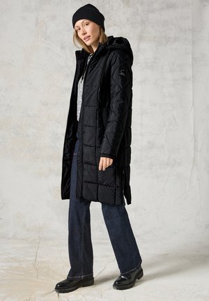 Abrigo largo acolchado negro con capucha, que presenta un cierre de cremallera, textura acolchada y bolsillos laterales. Combinado con denim oscuro y botas.
