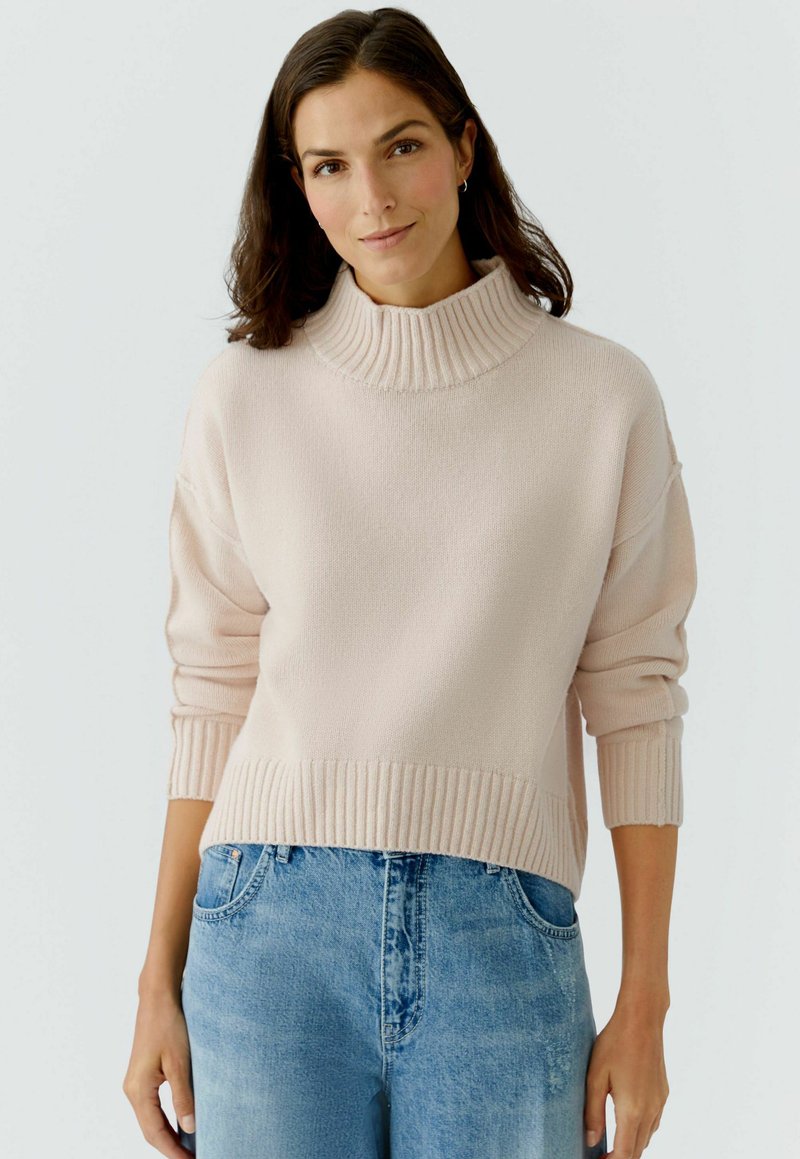 Helles rosa Strickpullover mit hohem geripptem Kragen und weiten Ärmeln, ausgestattet mit einem gerippten Saum und weicher Textur, kombiniert mit blauen Jeans.