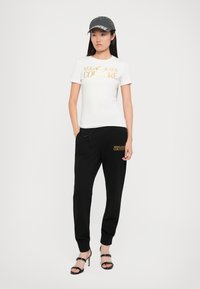 Camiseta blanca con el texto dorado "VERSACE JEANS COUTURE", combinada con pantalones deportivos negros que presentan el mismo logo. El modelo lleva una gorra gris y tacones negros.