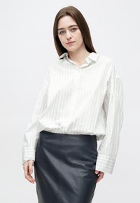 Młoda kobieta o prostych brązowych włosach, ubrana w długorękawową białą koszulę w paski i ciemną, dopasowaną spódnicę z wysokim stanem, stojąca na jednolitym tle.