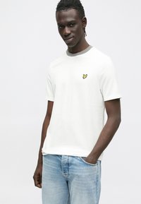 Bela majica s kratkimi rokavi, sivo okroglo ovratnico in majhnim rumenim vezenim logotipom na levi prsni strani. Nošena s svetlo modrimi jeans hlačami.