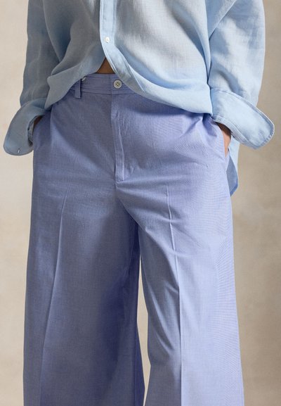 Άτομο που φοράει ανοιχτό γαλάζιο πουκάμισο με κουμπιά, φτιαγμένο μέσα σε ψηλόμεσο, φαρδύ παντελόνι σε λιλά απόχρωση, με τα χέρια στις τσέπες.