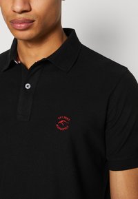 Schwarzes Poloshirt aus Baumwolle mit einer Drei-Knopf-Leiste. Mit rotem gesticktem Logo auf der Brust und gemustertem Detail im Kragen.
