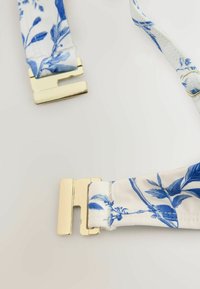 Bracelet en tissu blanc avec imprimé floral bleu et fermoir en métal doré partiellement ouvert sur un fond clair.