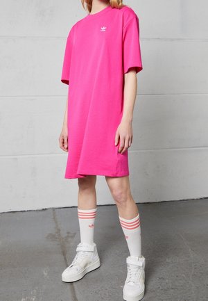 Osoba ubrana w luźną różową sukienkę t-shirtową Adidas, białe wysokie trampki oraz białe skarpetki z różowymi paskami, stojąca na szarej betonowej podłodze.