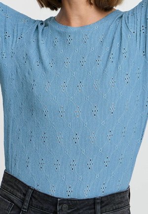 Vrouw draagt een lichtblauw, gestructureerd topje met korte mouwen en kleine uitsnijdingspatronen, ingestopt in een hoog uitgesneden zwarte spijkerbroek.