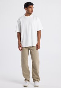 Joven de pie, vistiendo una camiseta blanca básica de gran tamaño, pantalones beige de pierna recta, zapatillas blancas y mirando hacia su derecha.