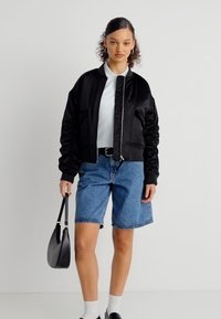 Νεαρή γυναίκα που φορά μαύρο σακάκι, λευκό πουκάμισο, μπλε τζιν σορτς, κρατά μια μαύρη τσάντα, στέκεται μπροστά σε απλό λευκό φόντο.