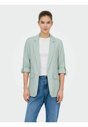 Femeie îmbrăcată într-un sacou verde deschis cu mâneci suflecate, cămașă albă și blugi albaștri, stând în fața unui fundal alb simplu.