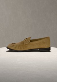 Mocassin en daim couleur fauve avec semelle noire, couture mocassin et bride décorative sur le dessus, présenté sur une surface et un fond neutres.