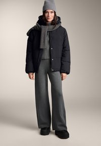 Jachetă neagră puf cu nasturi cu capse, eșarfă gri cu dungi și căciulă, bluză gri tricotată, pantaloni largi și pantofi negri pe un fundal neutru.