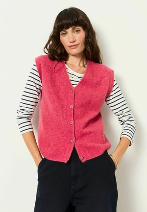 Frau mit gewelltem dunklem Haar trägt eine pinke, geknöpfte Weste über einem weiß-schwarzen, gestreiften Langarmshirt und steht mit den Händen in den Taschen.