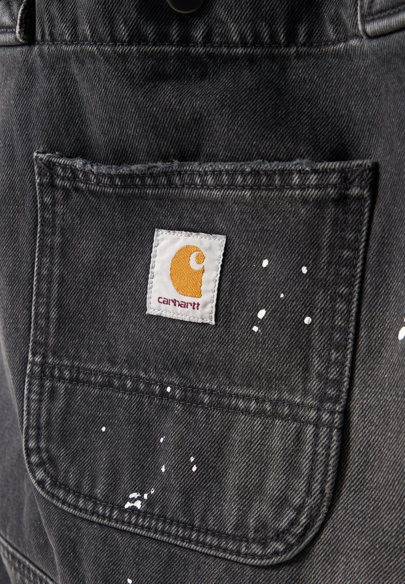 Schwarze Denimtasche mit weißen Farbspritzern und einem kleinen weißen Carhartt-Logo-Label, das nahe der oberen Mitte der Tasche angenäht ist.