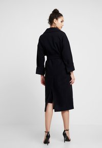 Schwarzer, taillierter Mantel mit Kragen, langen Ärmeln und einem Schlitz hinten. Weiche Textur und gerade Silhouette. Sichtbare hohe Schuhe.