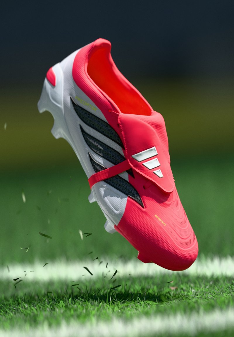 Scarpa da calcio rossa e bianca con strisce nere in aria sopra un campo d'erba con linea bianca, sollevando filo d'erba.