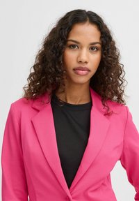 Fuchsiový růžový blazer s přizpůsobeným střihem, klopami a hladkým povrchem, nošený přes černý top. Kadeřavé vlasy rámují obličej.