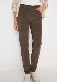 Bruine corduroy broek met een slim fit, voorzien van zijzakken en een knoopsluiting aan de voorkant. Gecombineerd met lichtgekleurde sneakers.