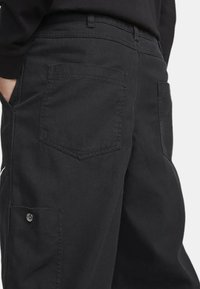 Mustat cargo-housut, joissa on teksturoitu kangas, useita taskuja ja suora lahje, sekä näkyvä nappi ja ommelkoristelu sivussa.