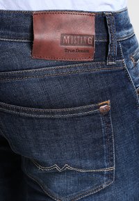 Džínsy z denimu s tmavomodrým umývaním, s koženou etiketou s nápisom "MUSTANG True Denim", oranžovým prešívaním a zadnými vreckami.
