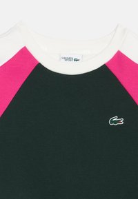 Тъмнозелена спортна риза Lacoste с бяла яка, розови ръкави raglan и малко зелено крокодилче на гърдите.